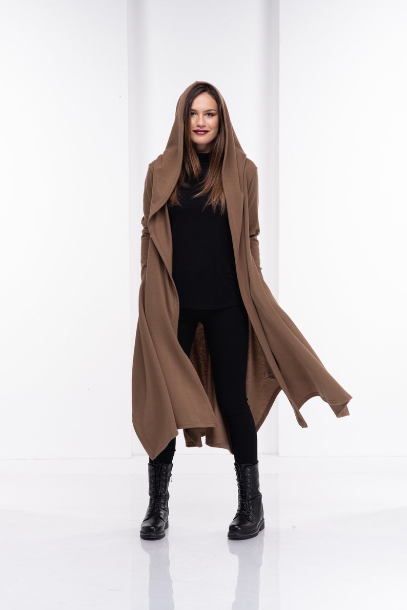 May include: A brown hooded cardigan with a long, flowing design. The cardigan is worn over a black turtleneck and black trousers. The person is wearing black boots.