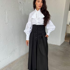 May include: A white button-down shirt with a large bow collar and long, puffy sleeves. Paired with a high-waisted, wide-leg black skirt with button detailing. The outfit is a modern take on classic formal wear.