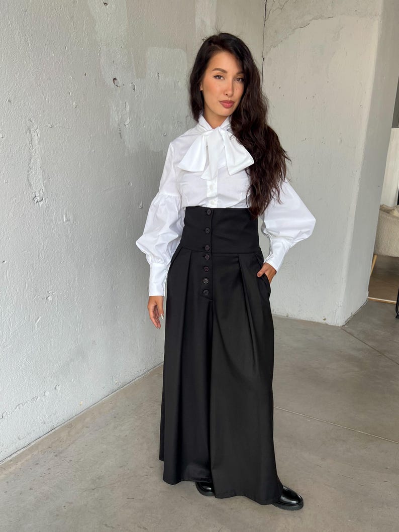 May include: A white button-down shirt with a large bow collar and long, puffy sleeves. Paired with a high-waisted, wide-leg black skirt with button detailing. The outfit is a modern take on classic formal wear.