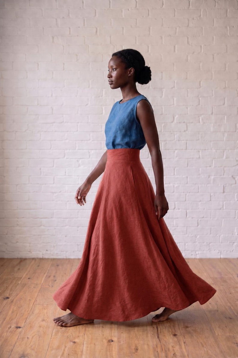 May include: A sleeveless blue top paired with a high-waisted, floor-length rust-colored skirt. The skirt features a wide waistband and a full, flowing silhouette that reaches the floor, crafted from a textured fabric with visible weave details.