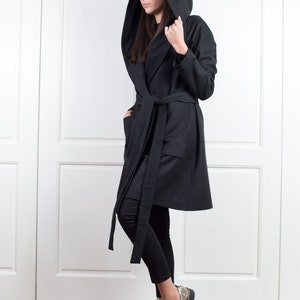 K&ouml;nnte beinhalten: Ein schwarzer Mantel mit Kapuze, Bindeg&uuml;rtel und Taschen. Der Mantel ist knielang und wird mit einer schwarzen Hose und metallisch-silbernen Schuhen getragen. Die Kapuze des Mantels ist hochgezogen.