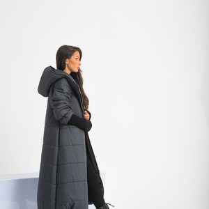 May include: A long, dark gray puffer coat with a hood. The coat has a quilted design and is worn by a person with long dark hair. The person is wearing black pants and black boots.
