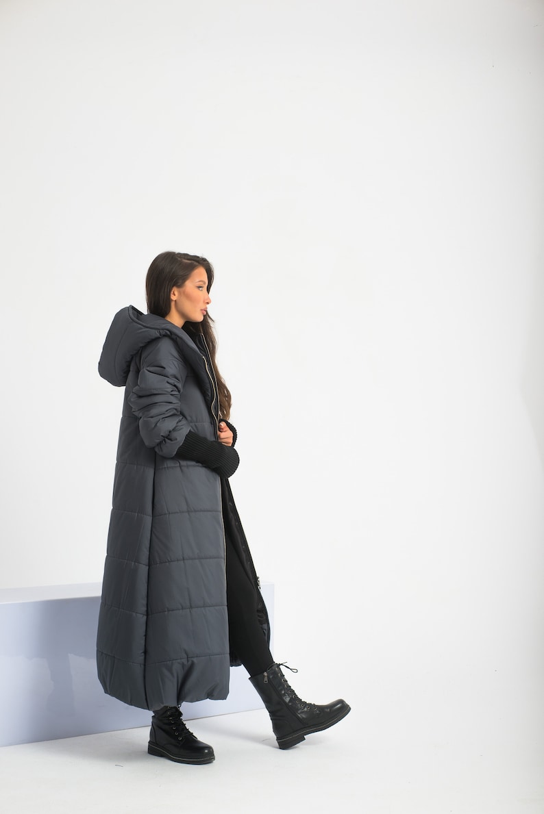 May include: A long, dark gray puffer coat with a hood. The coat has a quilted design and is worn by a person with long dark hair. The person is wearing black pants and black boots.