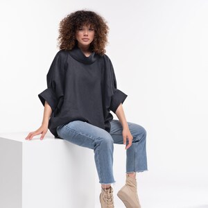 Puede incluir: Una mujer con una camisa negra de cuello alto y pantalones vaqueros azules se sienta en un cubo blanco. Lleva zapatillas de deporte beige de ca&ntilde;a alta.