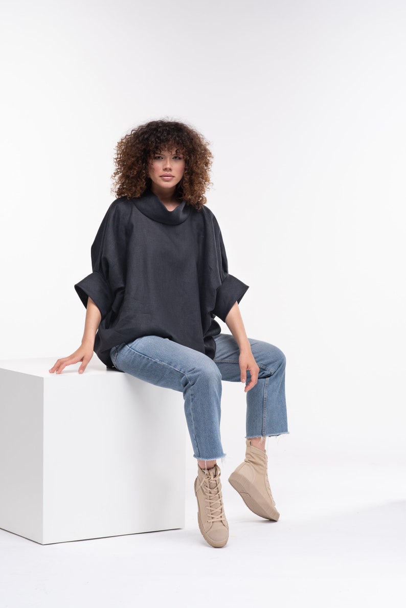 Puede incluir: Una mujer con una camisa negra de cuello alto y pantalones vaqueros azules se sienta en un cubo blanco. Lleva zapatillas de deporte beige de ca&ntilde;a alta.