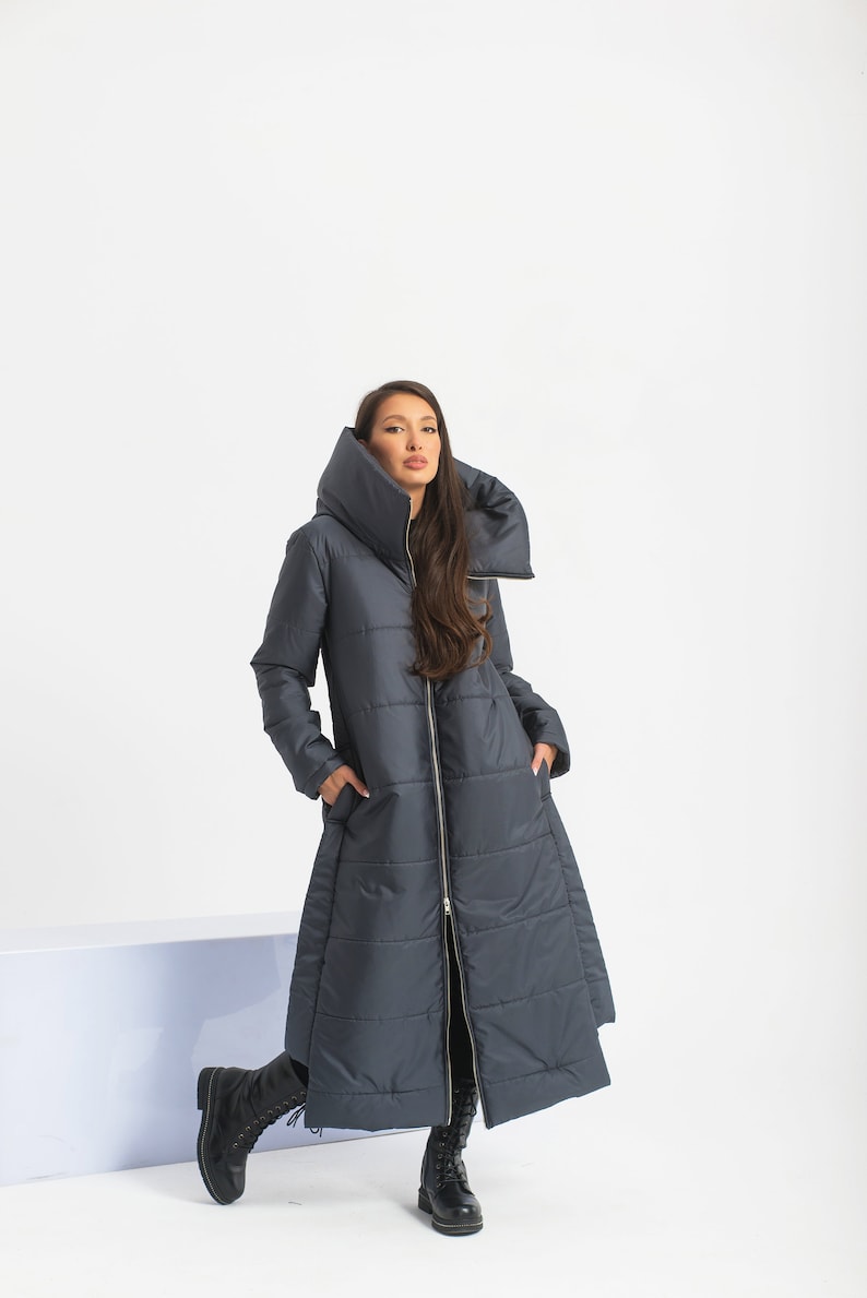 May include: A long, dark blue puffer coat with a large, folded collar. The coat has a zipper closure and is quilted throughout. The model is wearing black boots.