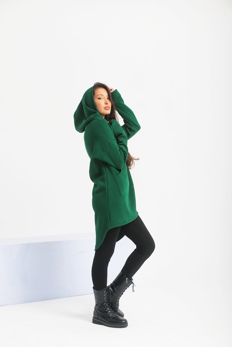 May include: A woman wearing a long, dark green hooded sweatshirt with a high-low hemline and black leggings. She is wearing black lace-up boots.