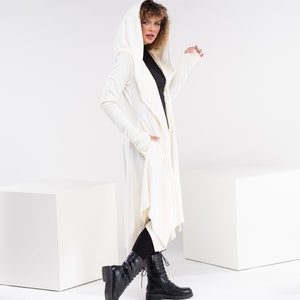 May include: A white hooded cardigan with long sleeves and a draped front. The cardigan is worn over a black turtleneck and black leggings. The model is wearing black lace-up boots.