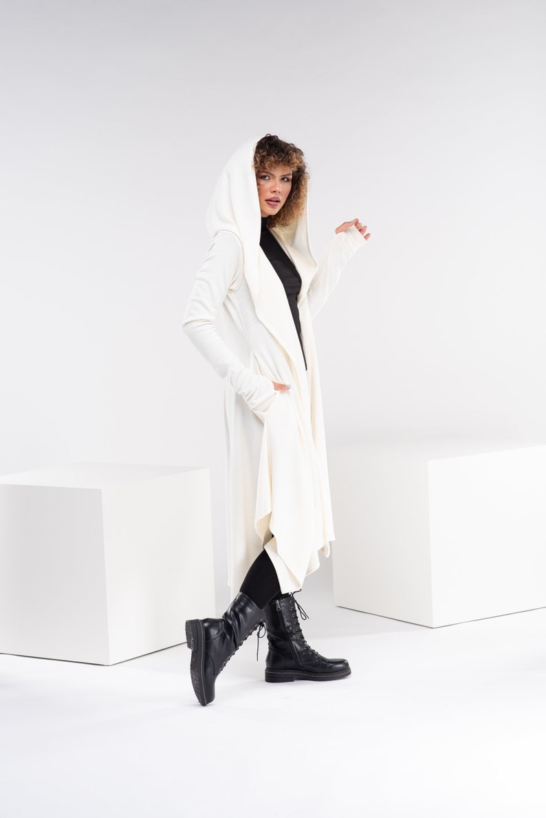 May include: A white hooded cardigan with long sleeves and a draped front. The cardigan is worn over a black turtleneck and black leggings. The model is wearing black lace-up boots.