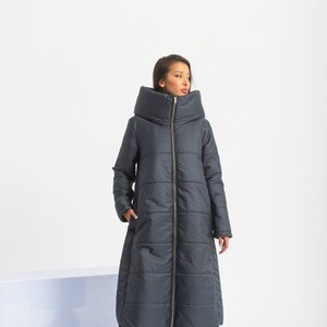 May include: A long, dark gray puffer coat with a large, folded collar. The coat has a zipper closure and a slightly flared hem.