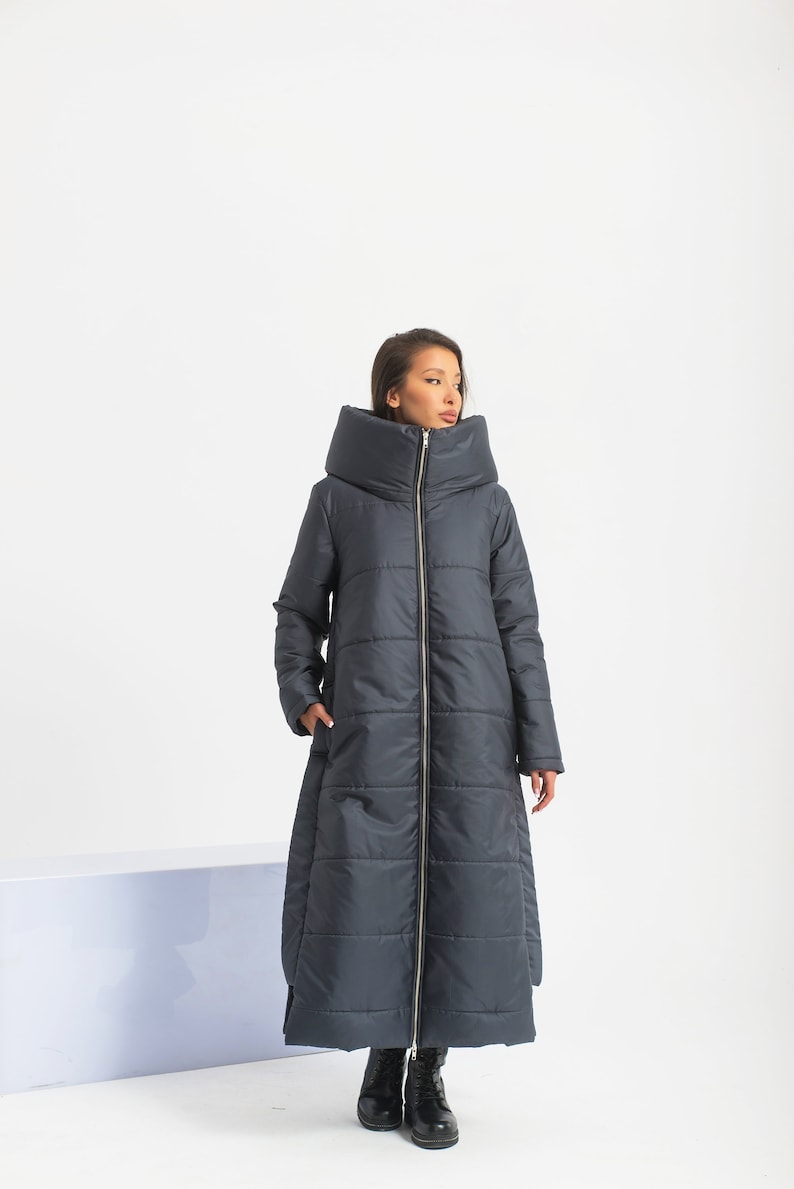 May include: A long, dark gray puffer coat with a large, folded collar. The coat has a zipper closure and a slightly flared hem.