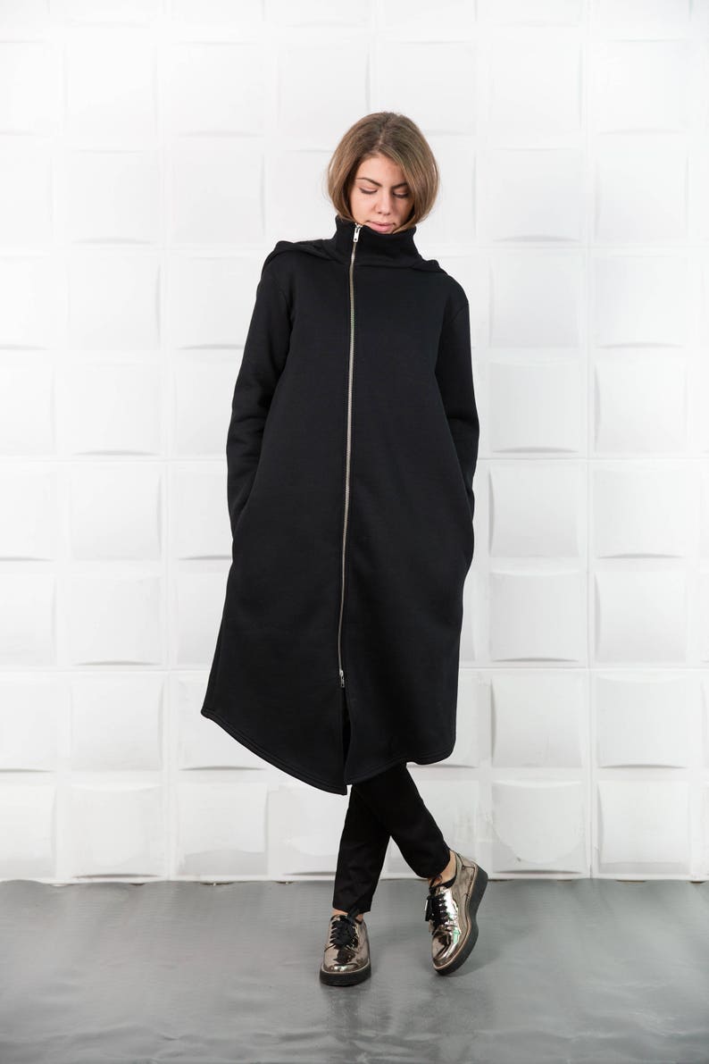 May include: A long black hooded jacket with a zipper closure. The jacket has large pockets on the sides and a relaxed fit. The model is wearing black pants and gold shoes.