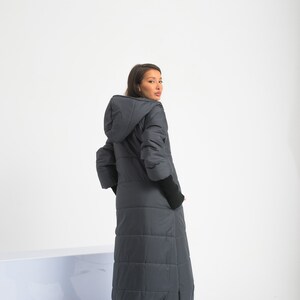 May include: A long, dark gray puffer coat with a hood. The coat has a quilted design and is worn by a person standing on a white surface.