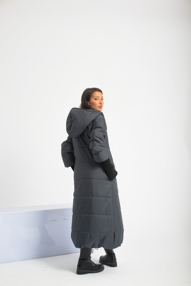 May include: A long, dark gray puffer coat with a hood. The coat has a quilted design and is worn by a person standing on a white surface.