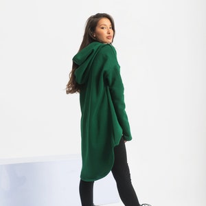 May include: A long, dark green hooded sweatshirt with a high, rounded neckline. The sweatshirt is made of a soft, comfortable fabric and has a relaxed fit. The model is wearing black leggings and black lace-up boots.