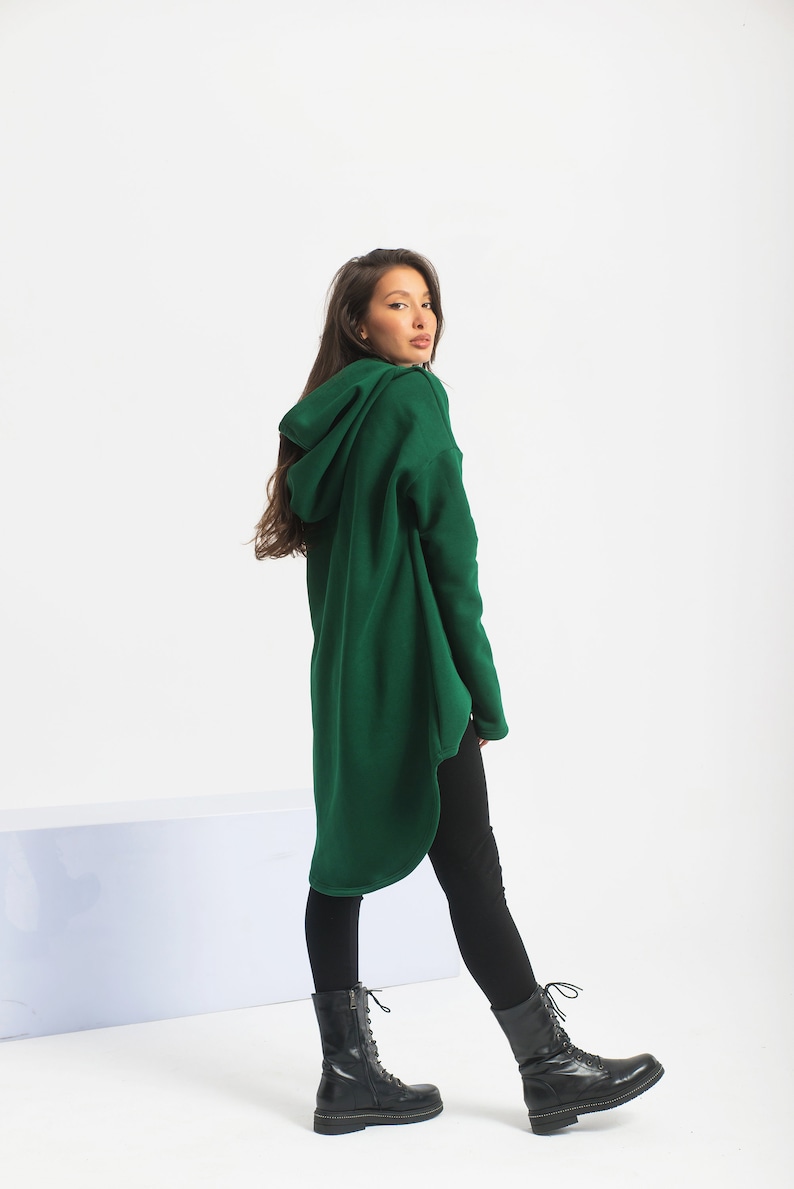 May include: A long, dark green hooded sweatshirt with a high, rounded neckline. The sweatshirt is made of a soft, comfortable fabric and has a relaxed fit. The model is wearing black leggings and black lace-up boots.