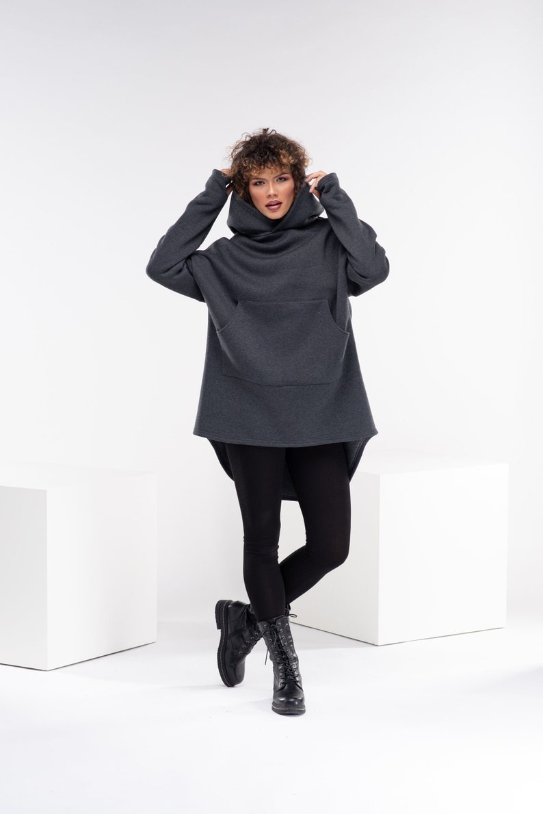 May include: A charcoal gray oversized hoodie with a high cowl neck and a kangaroo pocket. The hoodie is worn by a person with curly hair and black leggings. The person is wearing black combat boots.