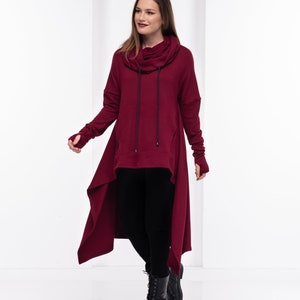 May include: A burgundy long-sleeved tunic with a cowl neck and a high-low hemline. The tunic has a drawstring at the neck and thumbholes at the cuffs. The model is wearing black leggings and black combat boots.