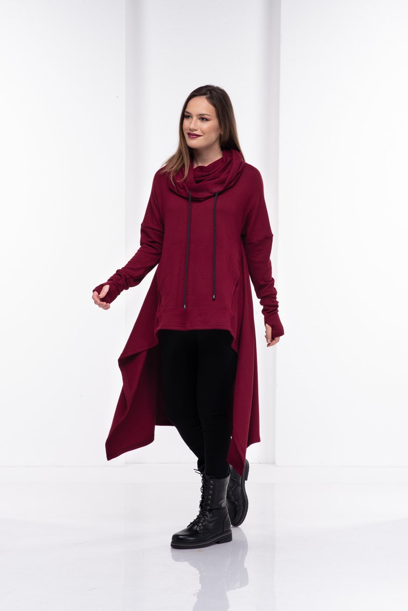 May include: A burgundy long-sleeved tunic with a cowl neck and a high-low hemline. The tunic has a drawstring at the neck and thumbholes at the cuffs. The model is wearing black leggings and black combat boots.