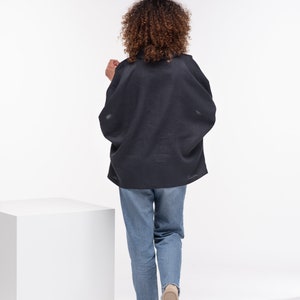 Puede incluir: Una persona lleva una blusa de lino gris oscuro de corte holgado y pantalones vaqueros azules. Se aleja de la c&aacute;mara, de espaldas al espectador.