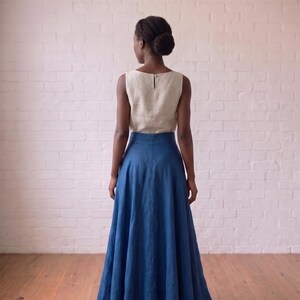May include: A sleeveless top in a light beige color with a round neckline and a small back button closure, paired with a floor-length, high-waisted skirt in a deep blue color. The ensemble features a simple, clean design with a flared skirt silhouette.