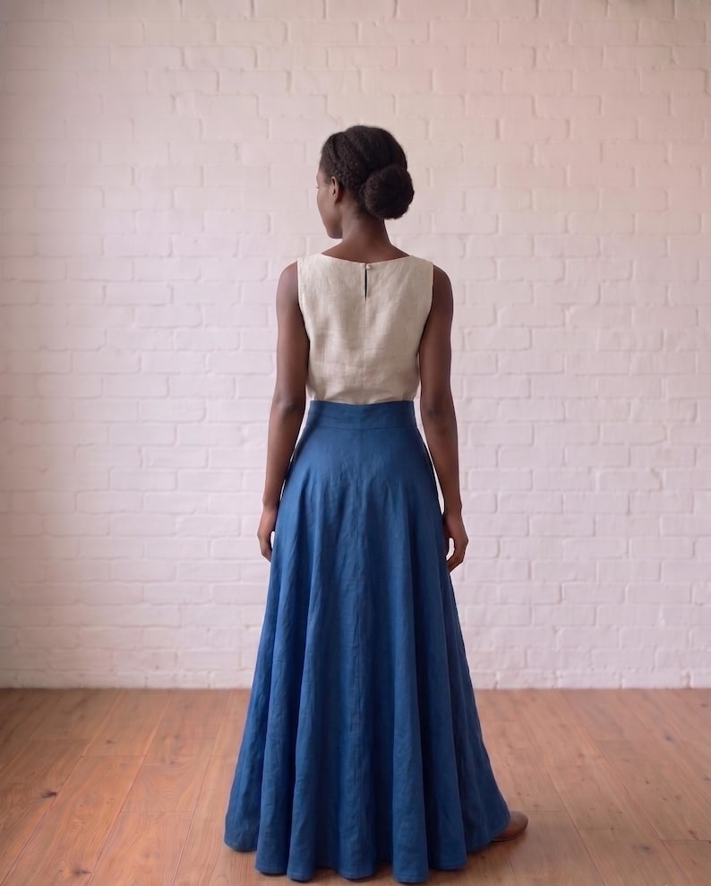 May include: A sleeveless top in a light beige color with a round neckline and a small back button closure, paired with a floor-length, high-waisted skirt in a deep blue color. The ensemble features a simple, clean design with a flared skirt silhouette.