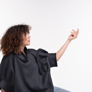 Puede incluir: Una mujer con una camisa negra de cuello alto y mangas largas de corte holgado. Lleva pantalones vaqueros azules y tiene el pelo rizado casta&ntilde;o.