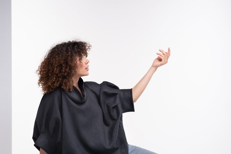 Puede incluir: Una mujer con una camisa negra de cuello alto y mangas largas de corte holgado. Lleva pantalones vaqueros azules y tiene el pelo rizado casta&ntilde;o.