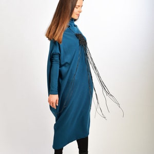 May include: A woman wearing a teal blue long-sleeved dress with a black fringe detail. She is wearing black boots.