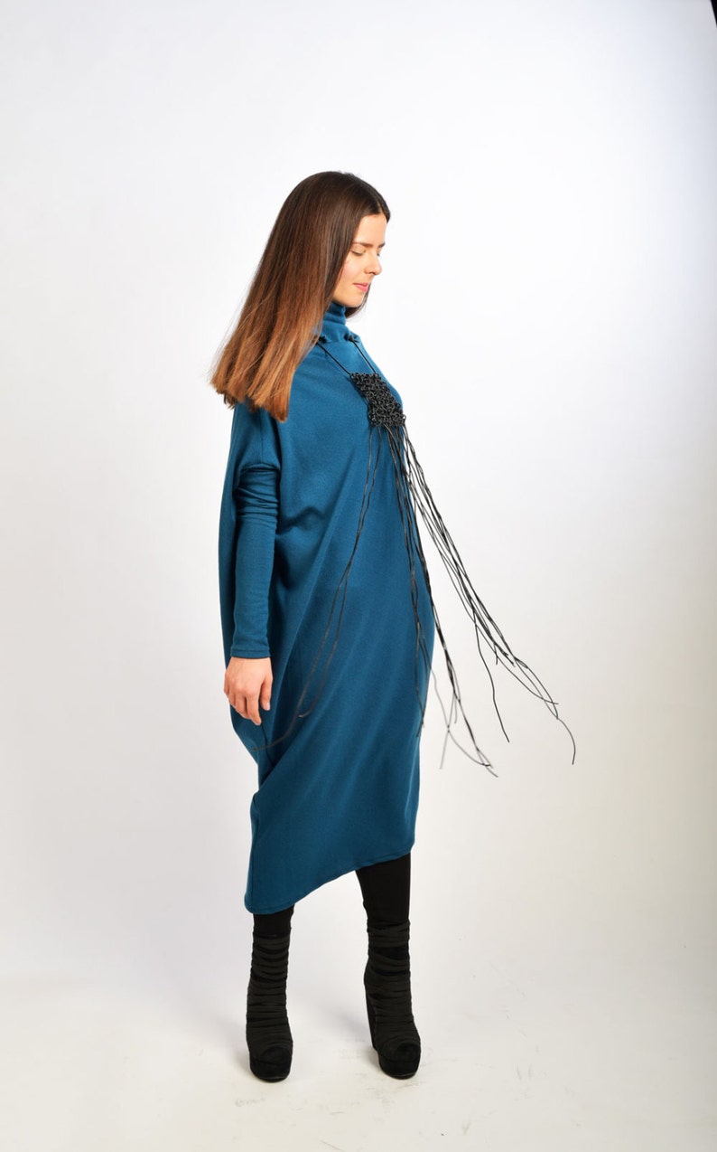 May include: A woman wearing a teal blue long-sleeved dress with a black fringe detail. She is wearing black boots.