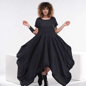 May include: A woman wearing a black asymmetrical linen dress with short sleeves. She is standing on a white platform with her feet in black boots.