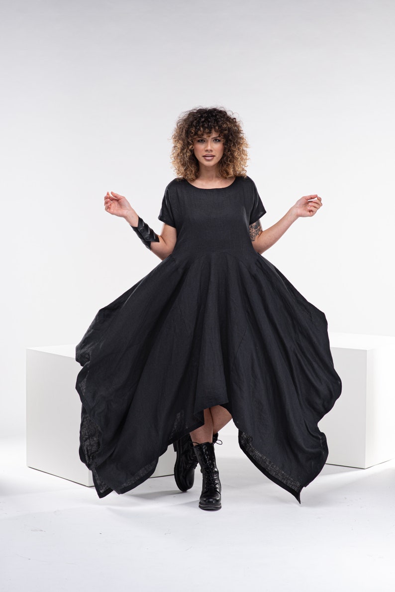 May include: A woman wearing a black asymmetrical linen dress with short sleeves. She is standing on a white platform with her feet in black boots.