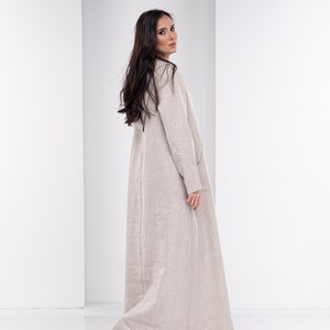 May include: A long, beige linen dress with a simple, relaxed fit. The dress has long sleeves and a V-neckline.