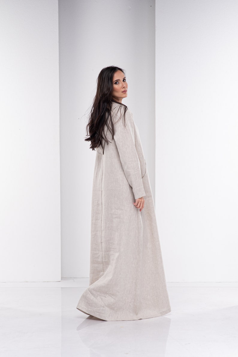May include: A long, beige linen dress with a simple, relaxed fit. The dress has long sleeves and a V-neckline.