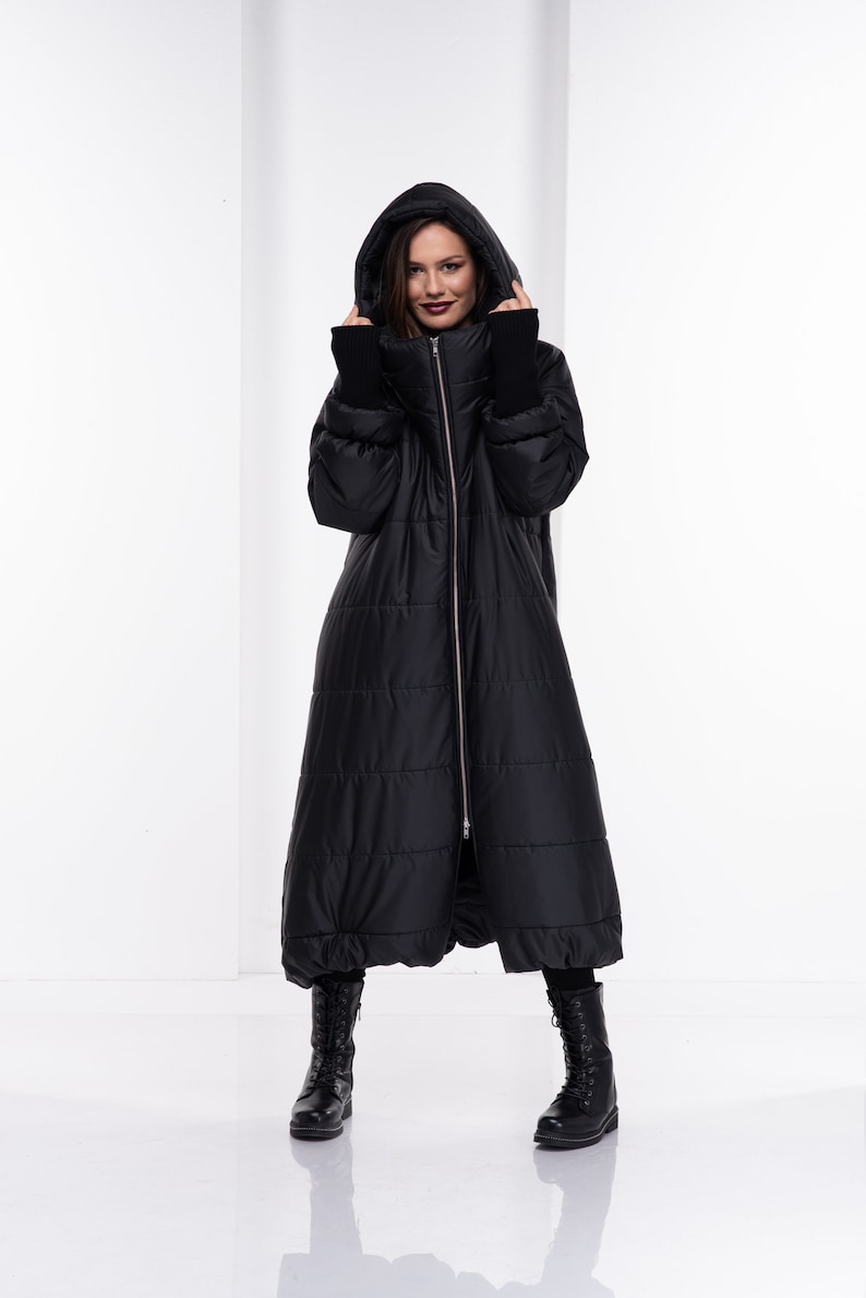 May include: A long black puffer coat with a hood and a zip closure. The coat has a quilted design and is worn by a person with black boots.