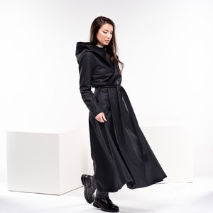 May include: A woman wearing a long black hooded coat with a belt. The coat has a flowing design and the woman is walking with her head down.