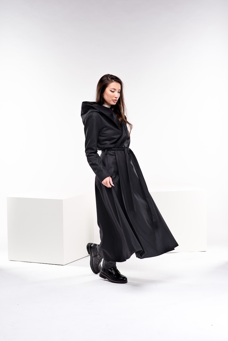May include: A woman wearing a long black hooded coat with a belt. The coat has a flowing design and the woman is walking with her head down.