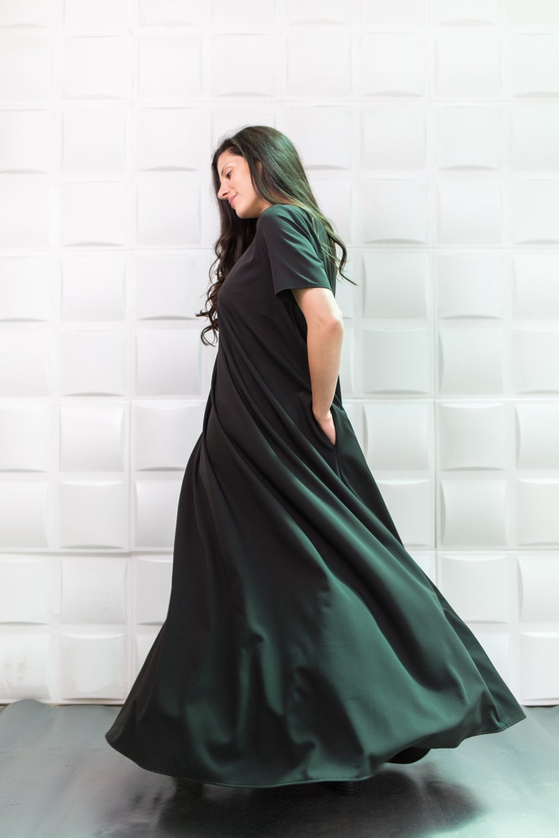 K&ouml;nnte beinhalten: Eine Frau tr&auml;gt ein langes, flie&szlig;endes schwarzes Kleid mit kurzen &Auml;rmeln. Das Kleid hat ein schlichtes Design und ist aus einem weichen, flie&szlig;enden Stoff gefertigt. Die Frau steht vor einer wei&szlig;en gekachelten Wand.
