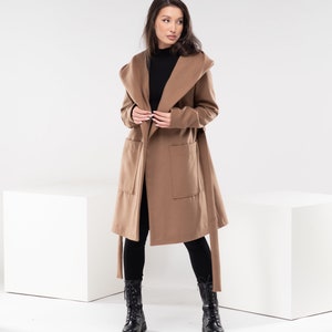 May include: A brown hooded wrap coat with a tie belt and two pockets. The coat is worn over a black turtleneck shirt and black pants. The person is wearing black lace-up boots.