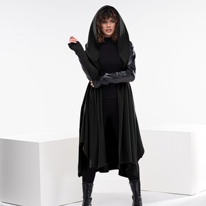 May include: A woman wearing a long black hooded cardigan with leather sleeves and black pants. She is standing on a white platform with a white background.