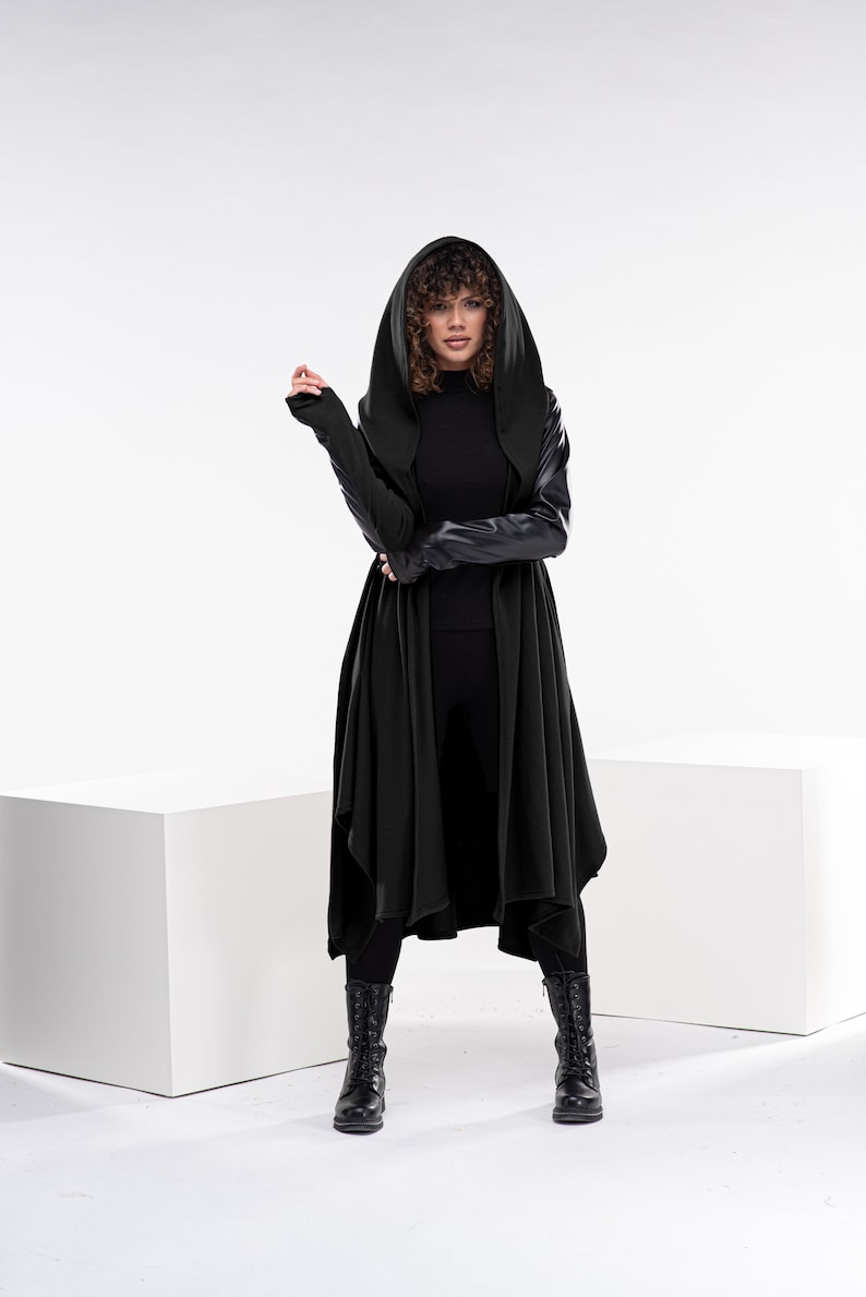May include: A woman wearing a long black hooded cardigan with leather sleeves and black pants. She is standing on a white platform with a white background.