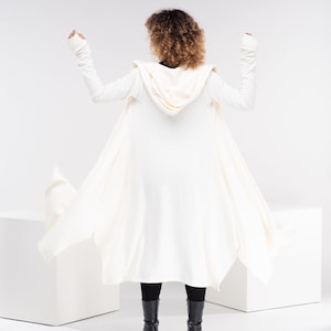 May include: A white hooded cardigan with long sleeves and a flowing design. The cardigan is worn by a person with curly hair, and the person is standing on a white surface.
