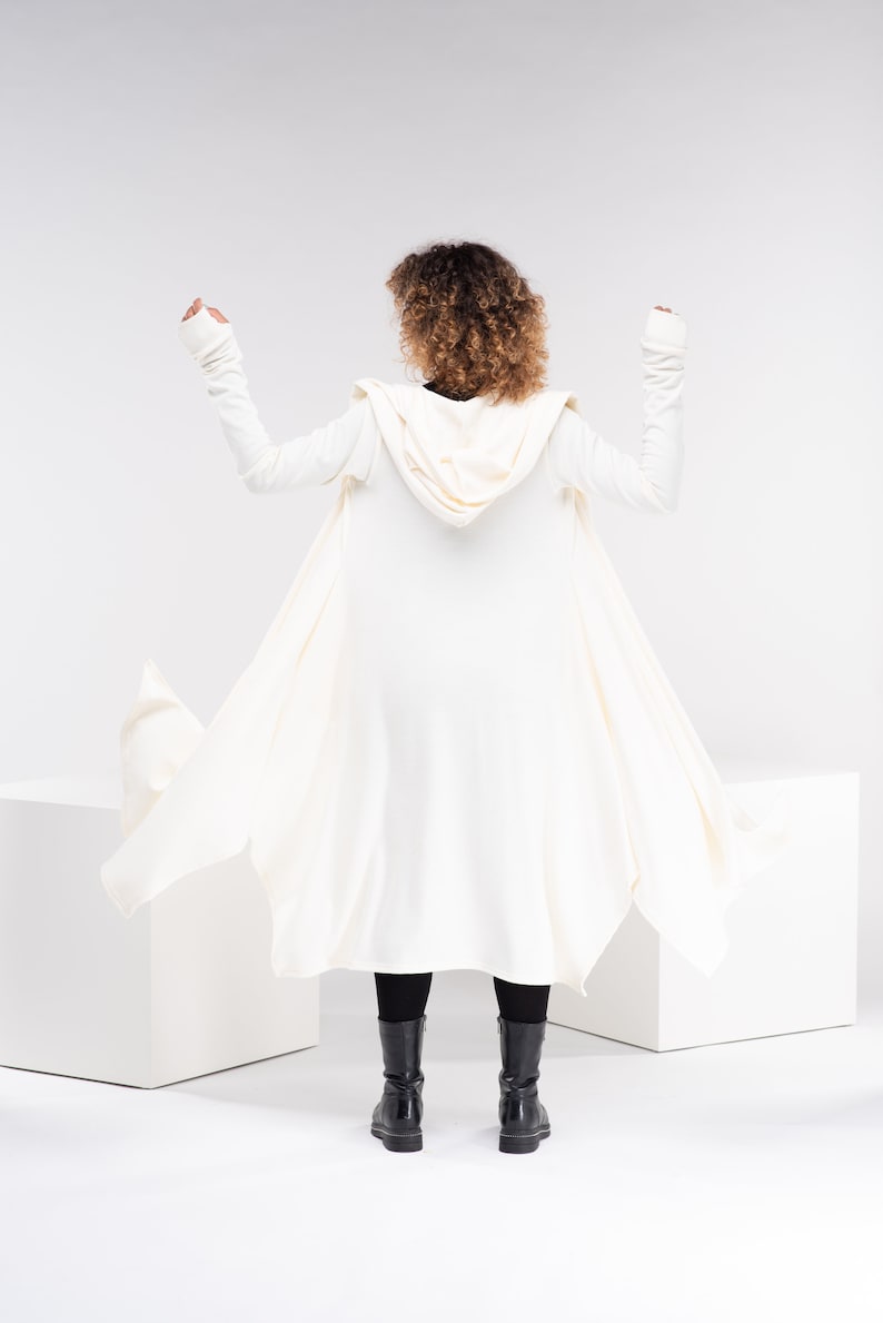 May include: A white hooded cardigan with long sleeves and a flowing design. The cardigan is worn by a person with curly hair, and the person is standing on a white surface.