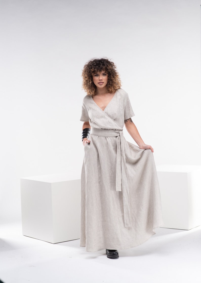 May include: A woman wearing a light beige linen wrap dress with short sleeves and a tie belt. The dress has a V-neckline and a flowing skirt. She is standing on a white background.