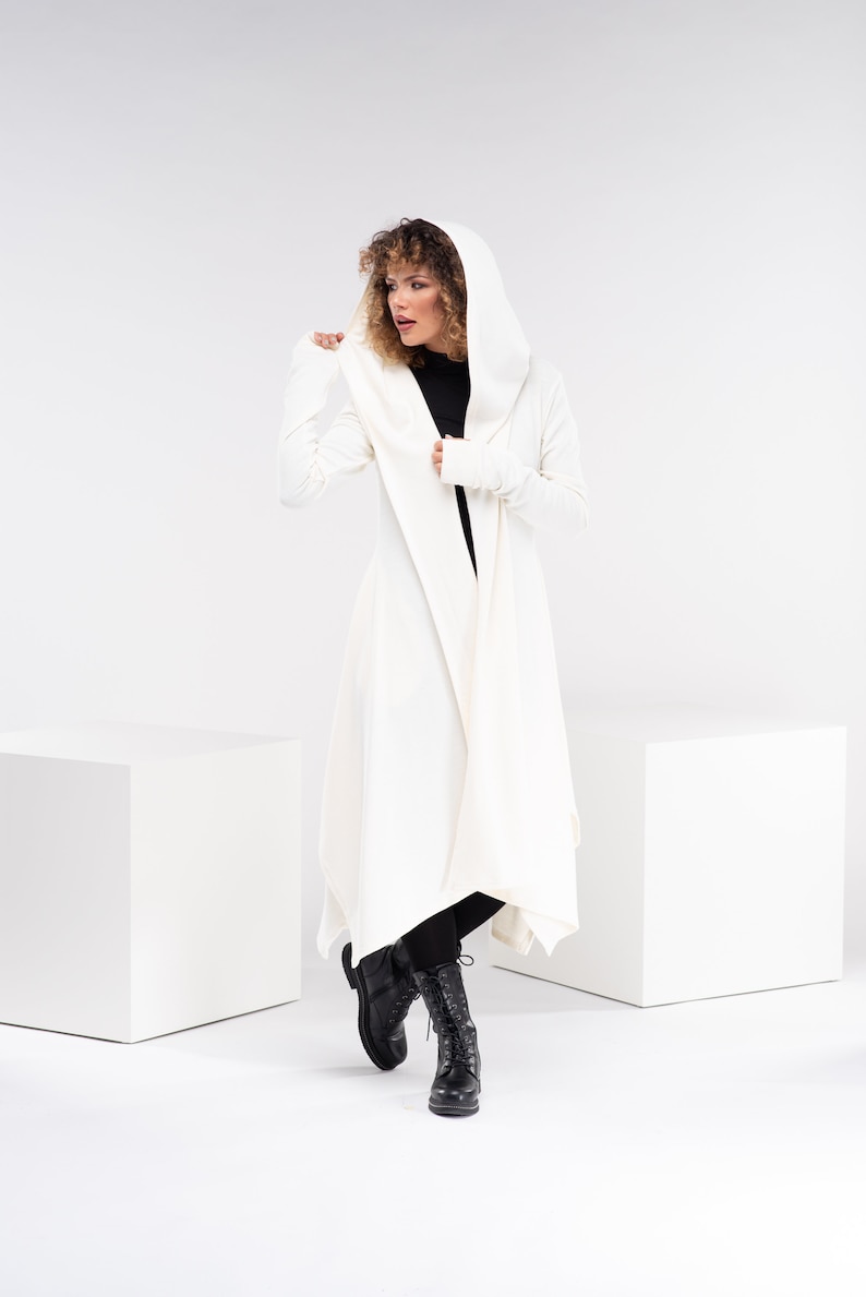 May include: A white hooded cardigan with long sleeves and a draped front. The cardigan is worn over a black top and black pants. The person is wearing black boots.