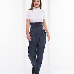 Pu&ograve; includere: Una donna indossa un top bianco a collo alto a maniche corte e pantaloni blu navy a vita alta. I pantaloni hanno una chiusura con bottoni e un design voluminoso unico. Le scarpe nere completano l'outfit, creando un look moderno ed elegante.