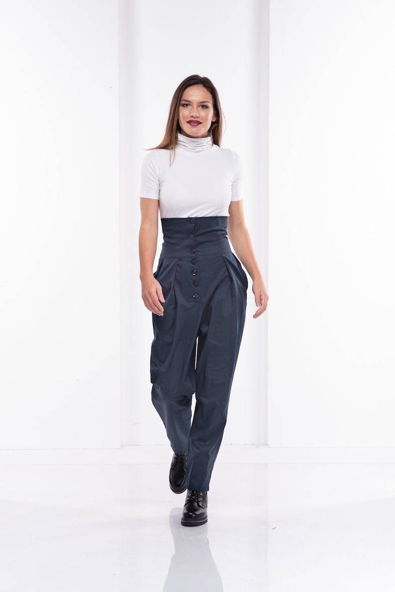 Pu&ograve; includere: Una donna indossa un top bianco a collo alto a maniche corte e pantaloni blu navy a vita alta. I pantaloni hanno una chiusura con bottoni e un design voluminoso unico. Le scarpe nere completano l'outfit, creando un look moderno ed elegante.