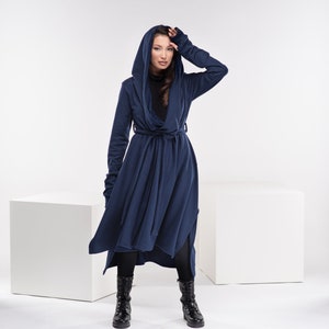 May include: A long, navy blue hooded cardigan with a tie belt. The cardigan has a draped, asymmetrical hemline and long sleeves with thumbholes. The model is wearing black leggings and black boots.