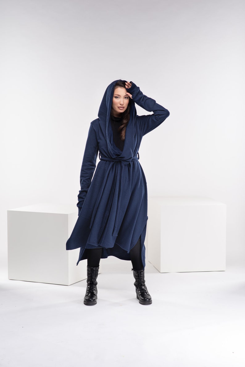 May include: A long, navy blue hooded cardigan with a tie belt. The cardigan has a draped, asymmetrical hemline and long sleeves with thumbholes. The model is wearing black leggings and black boots.