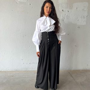 May include: A white blouse with a large bow and long, puffy sleeves paired with high-waisted black pants. The pants have a button-up closure and wide legs. The outfit is a stylish ensemble, suitable for various occasions.